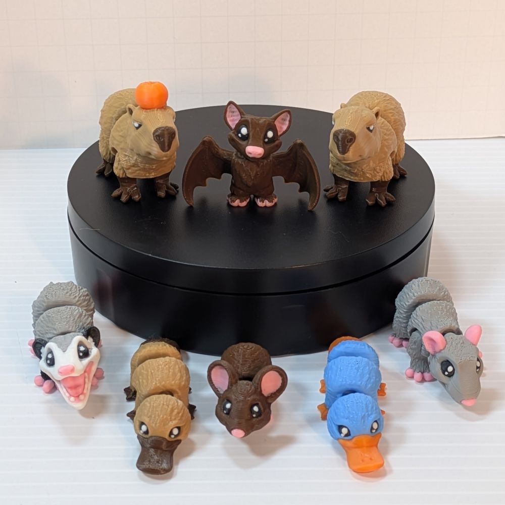 All of the current 3D printed fidget critters. On a stand are the capybara with fruit, bat, and capybara, and on the table beneath are the opossum, brown platypus, mouse, blue platypus, and rat.