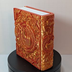 A 3D printed box that's shaped like a tome with fancy scrollwork and a ball of flame on the cover. It's made out of a shiny red/yellow filament, and it is turned to see the spine, which has the same fireball in the center.