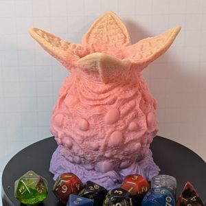 A 3D printed dice container that looks like an alien egg. It's lumpy, and there are four wedges open at the top, where you can drop in your dice. This one starts at a light purple at the bottom, fading up to pink and then a peach.