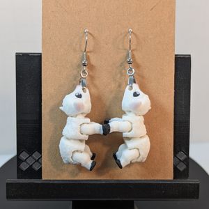 A pair of 3D printed earrings in the shape of lambs. They are white, with pink inside the ears and black hooves. They are hanging from silver earring hooks clasped into their mouths.