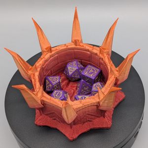 An orangish-red dice tray, that has bent spikes around the rim so it looks like a crown, filled with multi-colored dice.