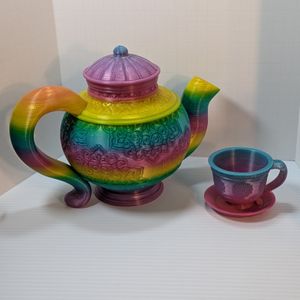 A 3D printed teapot, cup, and saucer. It has ornate work along the sides, and it's printed using a shiny rainbow gradient filament.
