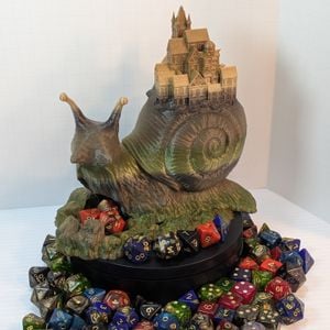A 3D printed dice tower in the shape of a snail, with a small town on the back of its shell. The snail is printed in a green and brown gradient, and it's surround by colorful TTRPG dice.
