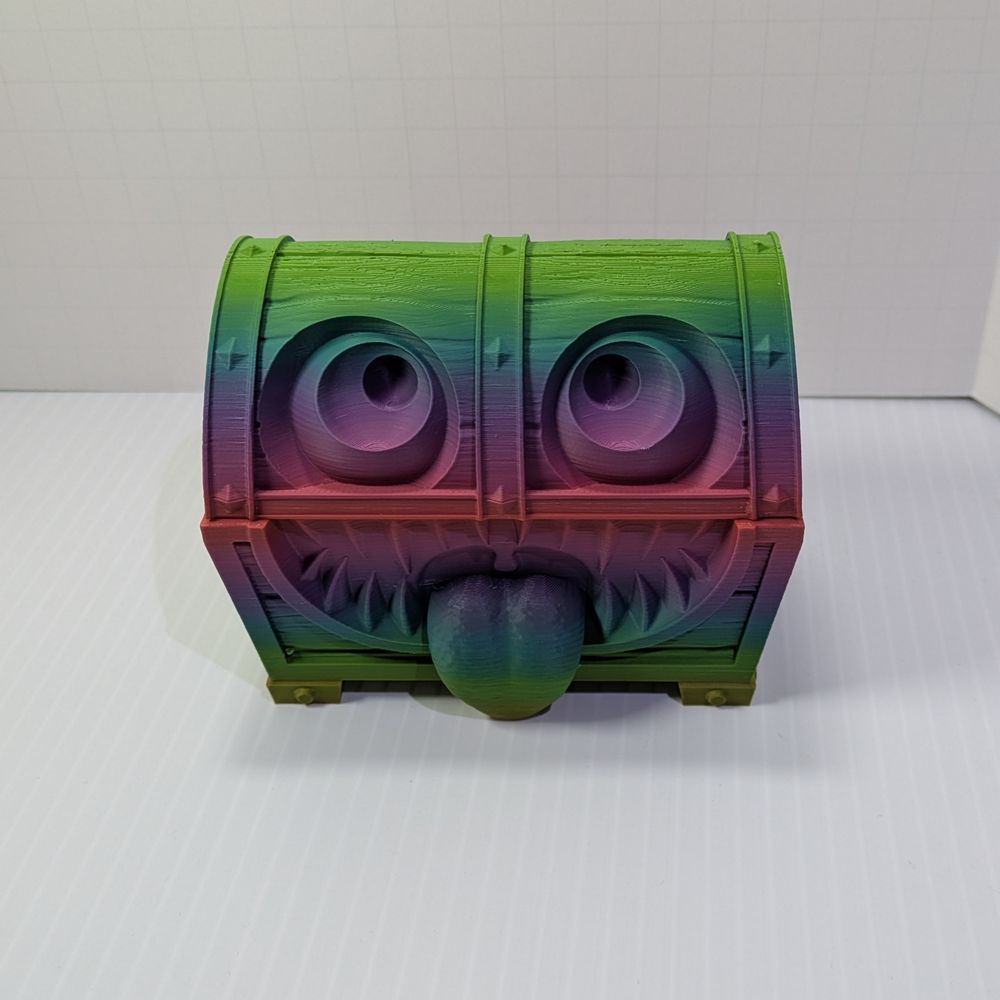 A 3D printed mimic, shaped like a treasure chest. The colors are a rainbow gradient, going from green at the bottom and top to purple in the center. It has large eyes, and a tongue sticking out of a toothy grin. It is very goofy looking.