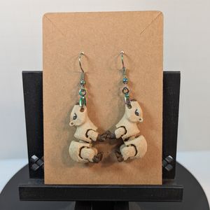 A pair of 3D printed earrings that look like capybaras. They are tan with brown feet and nose, an the eyes are in black and white. They are hanging on fishhook-style earring hooks on a carboard earring backer card.