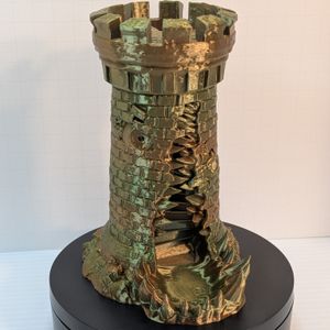 A 3D printed dice tower in the shape of a standard stone tower. This tower is in a shiny earth-tone filament, and has a large gaping maw up the side, in between two windows that look like eyes.