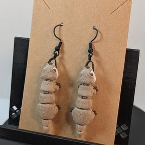 A pair of tan and white 3d printed river otter earrings, hanging on black fish hook style earring hooks from their mouths.