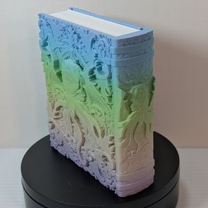 A dice box in the shape of a tome. It's printed in a muted gradient that goes from light blue, green, pink, to purple. On the cover is a face with many tentacles coming out of it. The spine is showing similar styling.