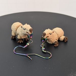 A pair of 3D printed earrings in the shape of a capybara. One is standing, the other is laying on its belly with their legs sticking out the sides.