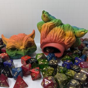 A 3D printed baby owlbear that is used to hold TTRPG dice. The main body is resting on its side, with the opening at the neck showing dice spilling out. The head is resting beside it, looking at the camera. It's surrounded by TTRPG dice.