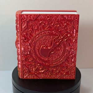 A 3D printed box that's shaped like a tome with fancy scrollwork and a ball of flame on the cover. It's made out of a shiny red/yellow filament, and has white "pages" in the center.
