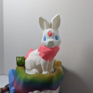 A closer shot of the bunny on top of the dice tower. It is white, with a pink tail and fur around the neck, blue eyes, and a pink gem in the forehead.