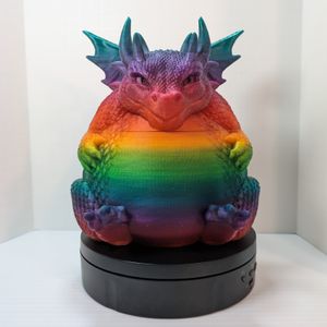 A 3D printed container of a very rotund dragon. It's sitting with is back feet sticking out from under its stomach, and its front paws grasping the sides of its belly. It has tiny wings and a mischievous grin on its face.  