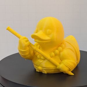 A yellow 3D printed rubber duck inspired piece dressed like a fantasy monk, with a bald head, simple clothing, and carrying a wooden staff across their chest in both hands.