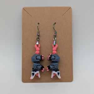 A pair of opossum earrings, grey and white with pink tails, their mouth open in a scream.