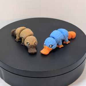 A light brown 3D printed platypus with darker brown bill and feet, next to a blue platypus with orange bill and feet. They are articulated at the neck, mid-body, sections of the tail, and at the legs.