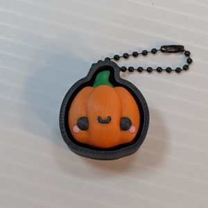 The 3D printed Pumpkin clicker, with a gall chain coming out of the top. The pumpkin is orange, with a green stem, pink cheeks, and black eyes and cute little smile. It's in a black case that gives it a black outline and hides the keyboard switch.