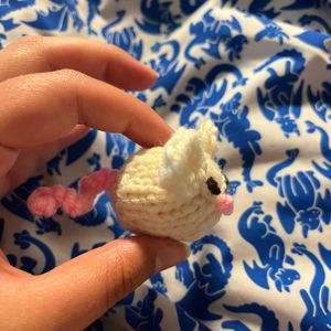 Anna, a white knitted mouse with a pink curly tail, a pink knot for a nose, brown knots for eyes, and big round white ears