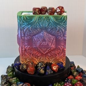 A 3D printed "tome", printed out of a shiny rainbow silk filament. On the front is a d20, showing the 20. There is a white insert for the "pages" and the entire piece is surrounded by colorful dice.