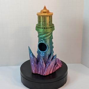 A 3D printed dice tower in the shape of a lighthouse, with a rocky base . It's printed in a very shiny rainbow filament. The back side of it shows where you drop the dice into, and how pointy the rocks on the base are.