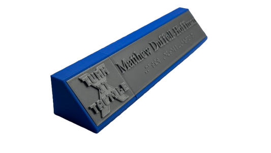 Angled view of the same tactile name display, highlighting the angled name plate face. The blue base supports a gray faceplate featuring the raised logo on the left and tactile name with braille on the right.