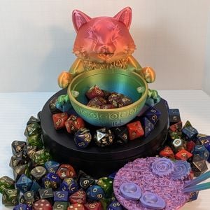 A 3d printed dice vault shaped like a chubby cat sitting behind a bow of ramen. The ramen and chopsticks are removed, showing a set of dice inside the bowl. It is surrounded by a colorful array of dice, and it rests on a black platform.