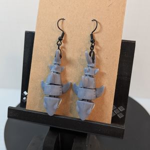 A pair of 3D printed earrings shaped like great white sharks. They're hanging by their tails from black fish hook shaped earring hooks from a carboard earring card.