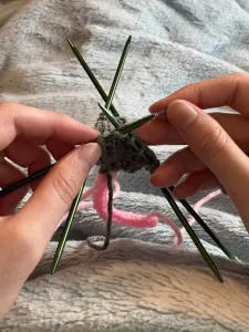 A photo of the artisan's hands knitting an opossum in the round. The pink tail is hanging down from the needles.