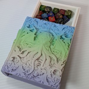 A dice box in the shape of a tome. It's printed in a muted gradient that goes from light blue, green, pink, to purple. On the cover is a face with many tentacles coming out of it.  The "pages" tray is partially slid out of the top, showing the rails.