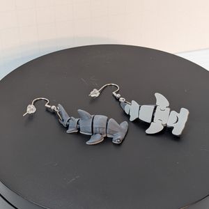 A 3d printed pair of earrings shaped like hammerhead sharks. They have silver-colored fish hook style earring hooks attached at the tails, and they've got a dark grey top and light grey underside. One is flipped so you can see a toothy little mouth.
