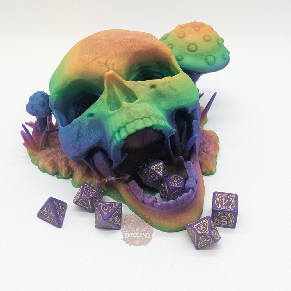A 3D printed skull in a rainbow filament. The jaw is open, and the eyes hollow, and it's a dice tower. It is resting on a base that looks like grass and mushrooms. There are dice around the tower, and inside the mouth where the dice tower empties out.