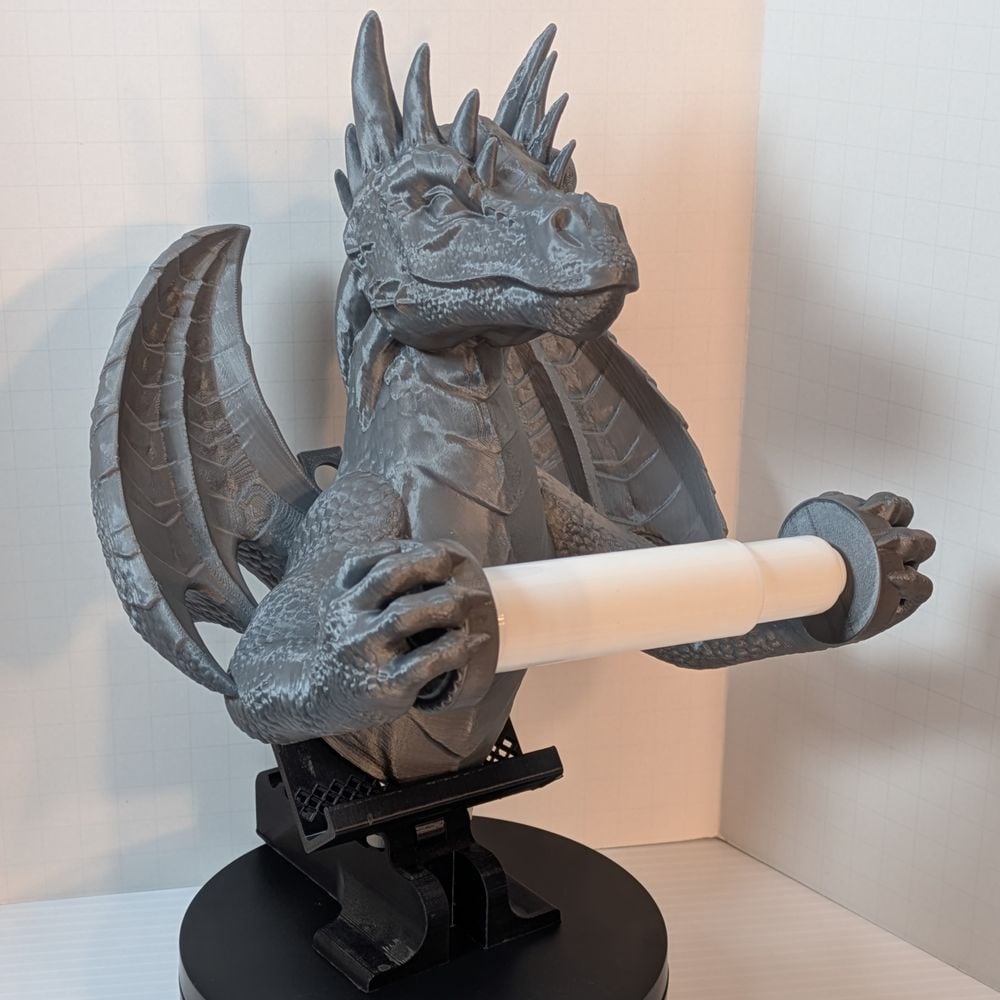 A 3D printed toilet paper holder in the shape of a dragon. It's a dull Steel color, and its front claws are holding the spindle for the toilet paper roll out away from its chest.