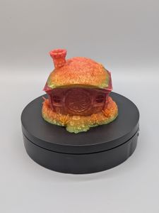 A 3D printed dice vault in the shape of the home of a halfling. It's got grass on the roof, a round chimney, and a round door. The top comes off to put the dice in. This one is a shiny rainbow gradient.