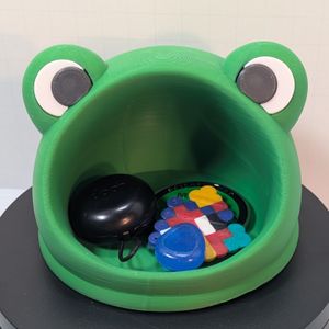 A green 3D printed catch all dish in the shape of a frog's head. The mouth is wide open for you to put stuff, and there are bumps at the top for the eyes. Inside the dish are some earbuds, a challenge coin, shiny rock, and a perler house.