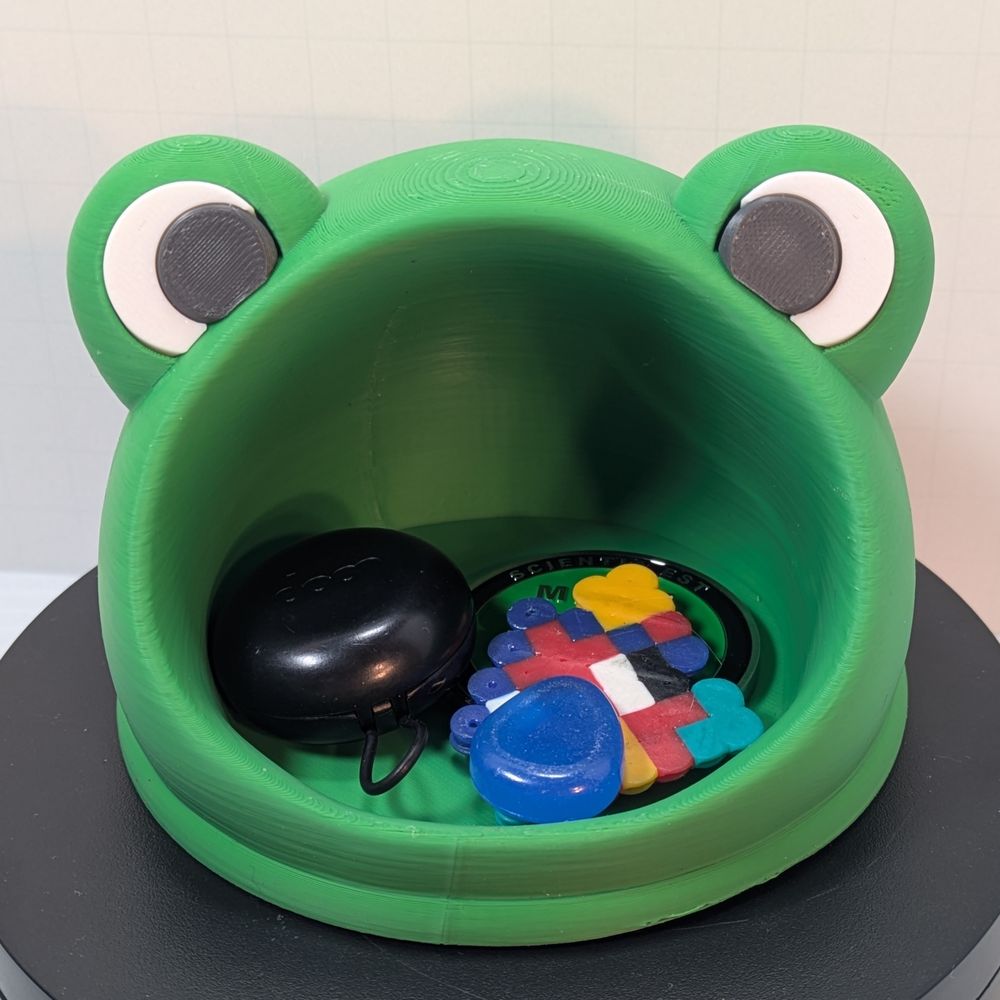 A green 3D printed catch all dish in the shape of a frog's head. The mouth is wide open for you to put stuff, and there are bumps at the top for the eyes. Inside the dish are some earbuds, a challenge coin, shiny rock, and a perler house.